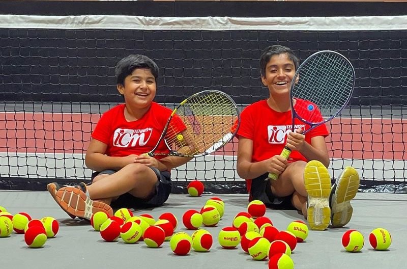 Two young ICM players sitting at the net with tennis balls around them.